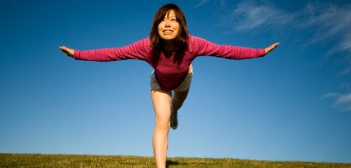 A woman in a pink sweater balances on one leg with her arms outstretched, smiling against a blue sky and green grass.