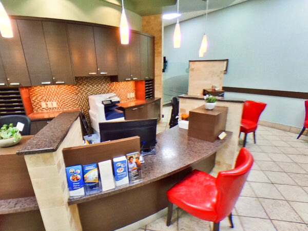A welcoming dental office reception area featuring a modern counter, red chairs, and information displays. The space is well-lit and organized, creating a friendly atmosphere for patients. A friendly blue 123Dentist Smiley adds a cheerful touch to the environment.