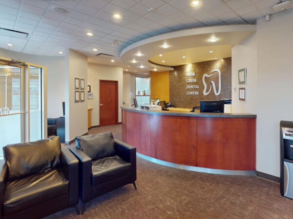 A modern dental office reception area featuring a wooden front desk, comfortable chairs, and warm lighting. The space has minimalistic decor, with a large tooth logo on the wall and a water cooler in the corner, creating a welcoming environment for patients.