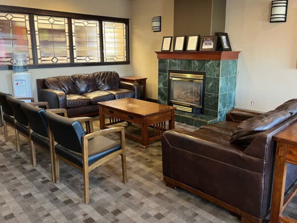 A cozy dental office waiting area featuring comfortable seating, a fireplace, and decorative elements. Natural light comes through the windows, creating an inviting atmosphere for patients.
