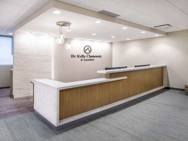 Reception area of a medical office featuring a modern front desk, neutral tones, and a company logo on the wall. Natural light enters through a window, creating a welcoming atmosphere.
