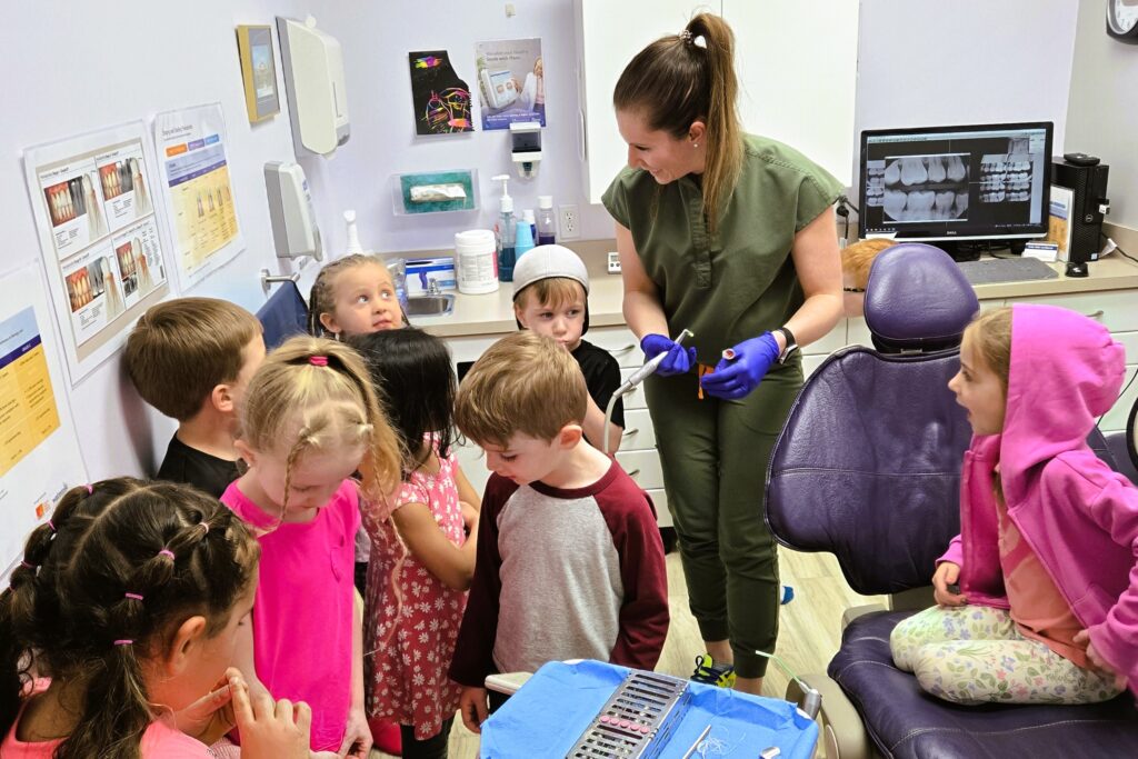 A dental hygienist engages a group of children in a pediatric dental office, demonstrating tools and fostering a friendly atmosphere. Some kids are gathered around, while others sit nearby, showing curiosity and excitement about their dental health. The 123Dentist Smiley adds a cheerful touch to the scene.