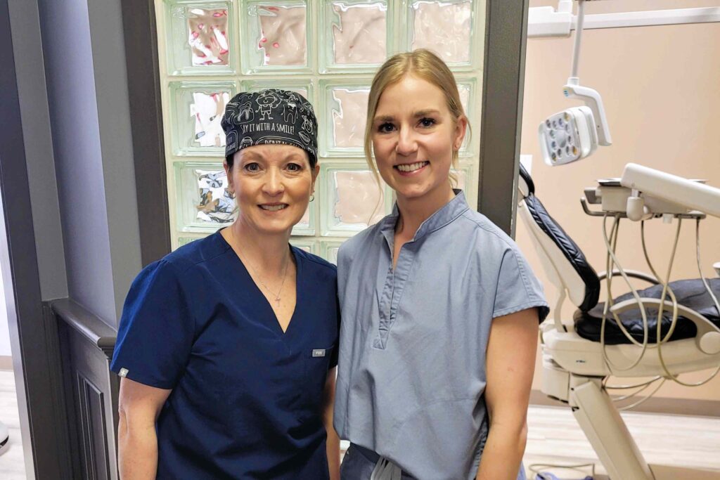 A dental professional wearing scrubs and a cap smiles alongside a colleague in a dental office. In the background, dental equipment is visible, creating a welcoming atmosphere.
