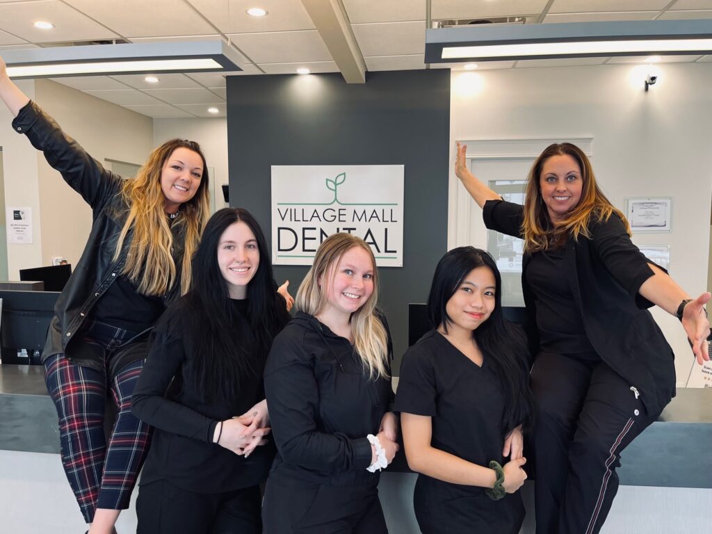 A cheerful dental team poses at the Village Mall Dental office, with staff members dressed in black. They are smiling and showcasing a friendly atmosphere, welcoming patients. The setting is bright and professional, conveying a sense of care and community.
