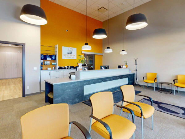 A modern waiting room featuring several orange chairs, a reception desk with a light-colored counter, and decorative wall accents. The space is brightly lit with pendant lights hanging from the ceiling, creating a welcoming atmosphere.