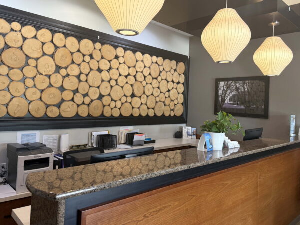 A reception area featuring a wooden front desk with a dark granite countertop. Above the desk is a decorative wall made of round wooden logs, and there are three pendant lights hanging from the ceiling. A small plant and various office supplies are visible on the counter.