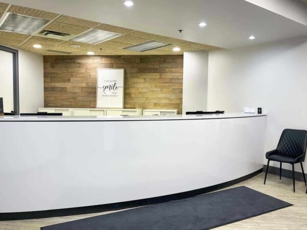 A modern reception area featuring a curved white desk, wooden accent wall, and two black chairs beside a black mat. The space is well-lit with ceiling lights, creating a welcoming atmosphere.