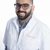 A smiling individual wearing glasses and a white lab coat stands against a plain background. The person has a short beard and is dressed in a checked shirt beneath the coat, presenting a professional appearance.