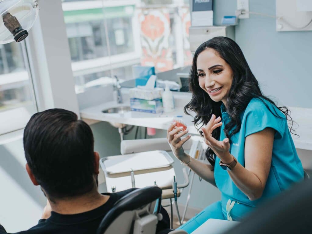 A dental professional in a blue outfit is engaging in a friendly conversation with a patient seated in a dental chair. The bright and inviting clinic features natural light, creating a warm atmosphere, while tools and equipment are visible in the background. The 123Dentist Smiley adds a cheerful touch.