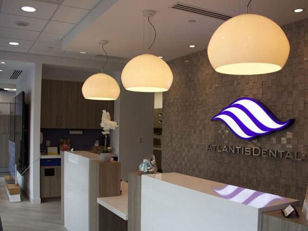 Modern dental office reception area with a sleek design, featuring a large logo on the wall, pendant lights, and white desks.