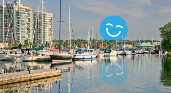Une scène sereine de marina avec plusieurs voiliers amarrés dans des eaux calmes, avec des bâtiments modernes en arrière-plan et un joyeux visage souriant bleu superposé.