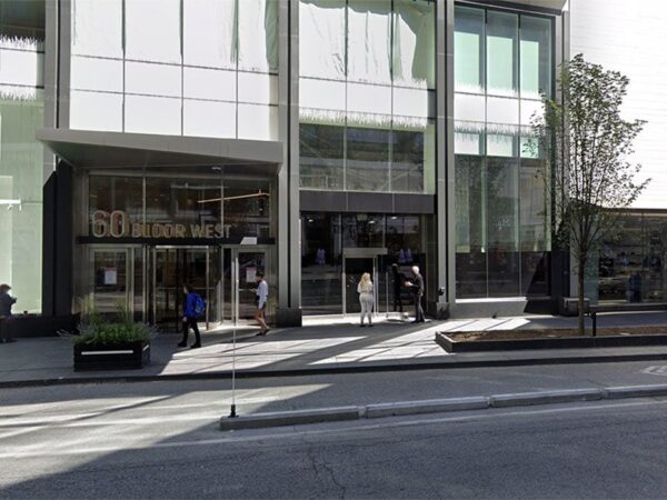 A modern building with large glass windows features an entrance with people walking in and out. The exterior includes a small tree and planters along the sidewalk, while the street is lightly trafficked.