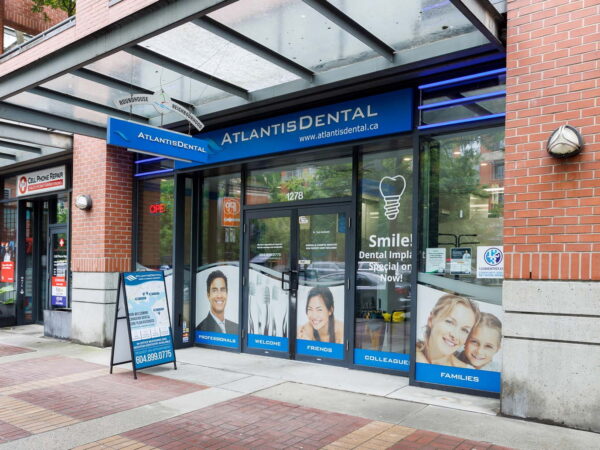 Exterior of Atlantis Dental clinic featuring large glass doors, blue signage, and advertisements displaying smiling individuals. A sidewalk sandwich board promotes dental services, with a modern brick building in the background.