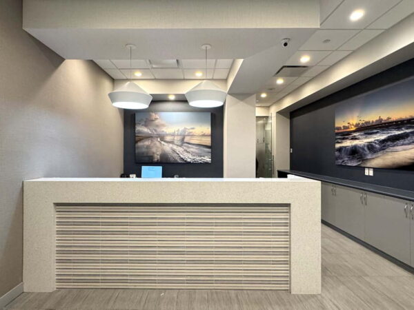 A modern reception area featuring a sleek white desk with horizontal lines, pendant lighting, and large wall-mounted landscape photographs of a beach scene. The walls are painted in neutral tones, creating a bright and inviting atmosphere.