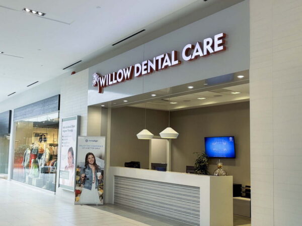 A modern dental care office with a reception area featuring the sign "Willow Dental Care." The space is brightly lit and includes a small seating area and a monitor. Promotional materials are displayed nearby.