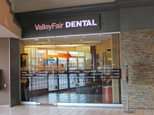 The image shows the entrance of ValleyFair Dental, featuring a glass façade with the dental office name prominently displayed. Inside, there are bright lights and modern furnishings. The setting is a shopping mall environment.
