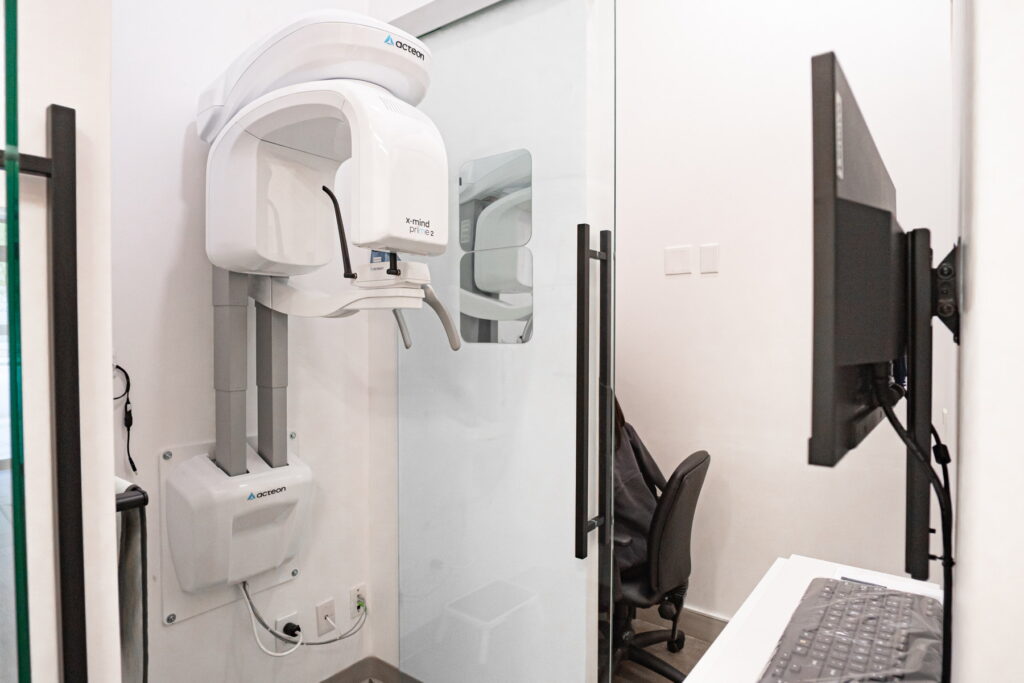 A dental imaging room featuring a white cone beam CT scanner against a glass wall, with a computer monitor and office chair visible. The space is modern and designed for dental diagnostics.