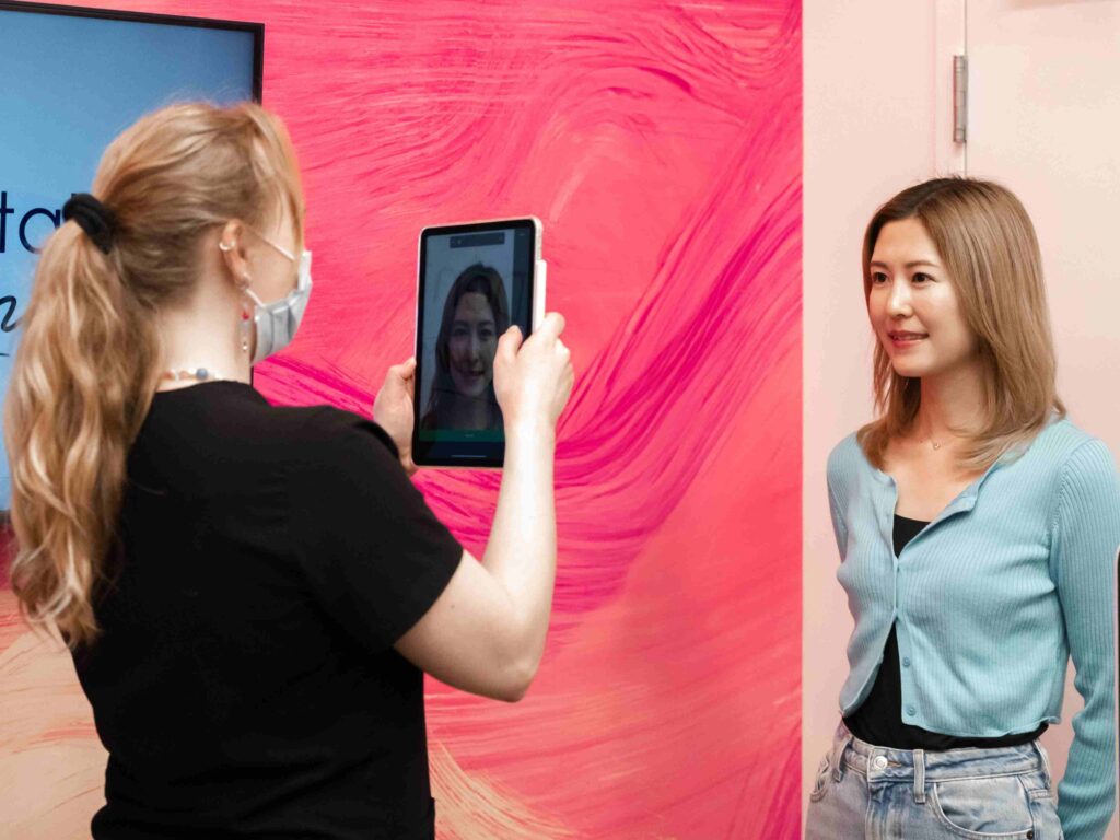A woman is having her photo taken by another woman using a tablet in front of a colorful, swirling pink backdrop. The scene reflects a friendly atmosphere, possibly in a dental office setting. The 123Dentist Smiley is nearby, adding a cheerful touch.