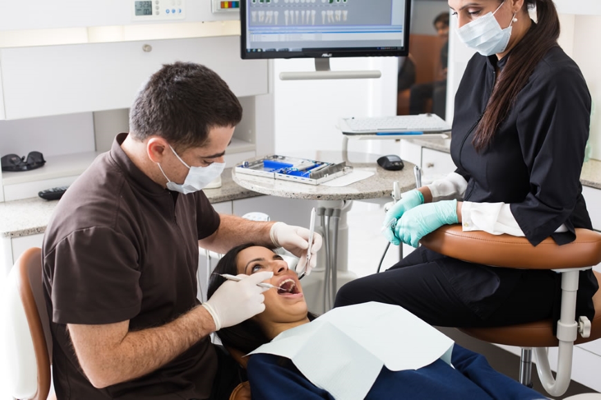 A dentist is examining a patient seated in a dental chair, while an assistant stands beside them, both wearing masks and gloves. The patient appears relaxed with a light dental bib. A dental monitor displays information in the background. The friendly 123Dentist Smiley adds a cheerful touch to the scene.