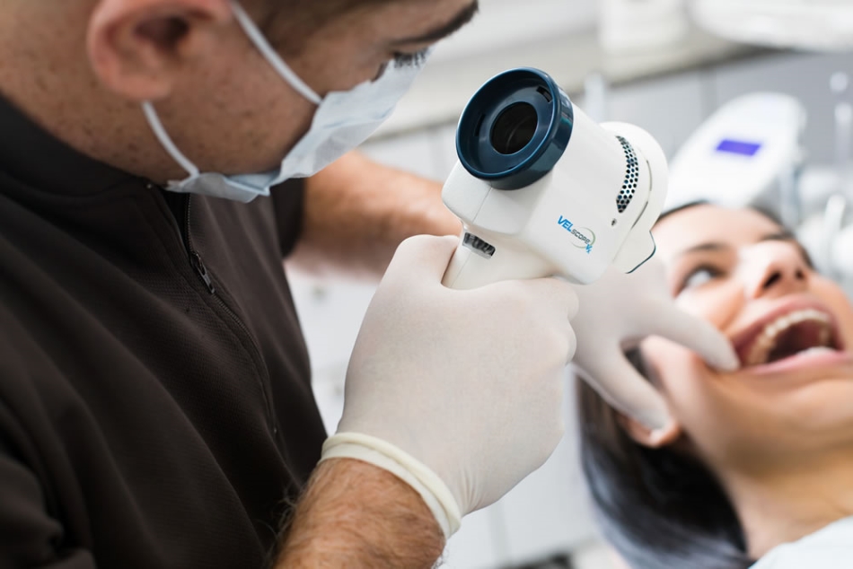 A dentist, wearing a mask and gloves, examines a patient's teeth using a dental camera. The patient is smiling, showing comfort during the procedure. The scene reflects a positive dental care experience. The friendly 123Dentist Smiley can be seen nearby, adding a cheerful touch.