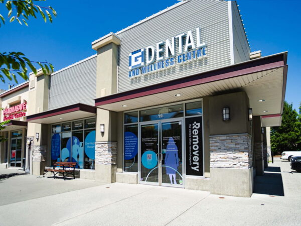 A modern dental clinic with glass windows showcasing the name "IQ Dental." The building features a light gray exterior with stone accents, surrounded by greenery and parked cars, indicating a welcoming, professional environment for dental care.