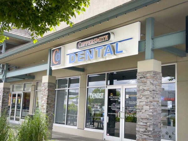 A dental office storefront featuring a prominently displayed sign with the word "DENTAL" in blue letters. The entrance has large glass doors and stone columns, surrounded by other commercial storefronts.