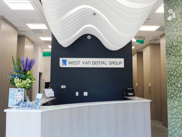 A modern dental office reception area featuring a sleek white front desk, a dark wall displaying the "West Van Dental Group" sign, and decorative plants. The design is contemporary with wave-like ceiling features and green textured wall accents.