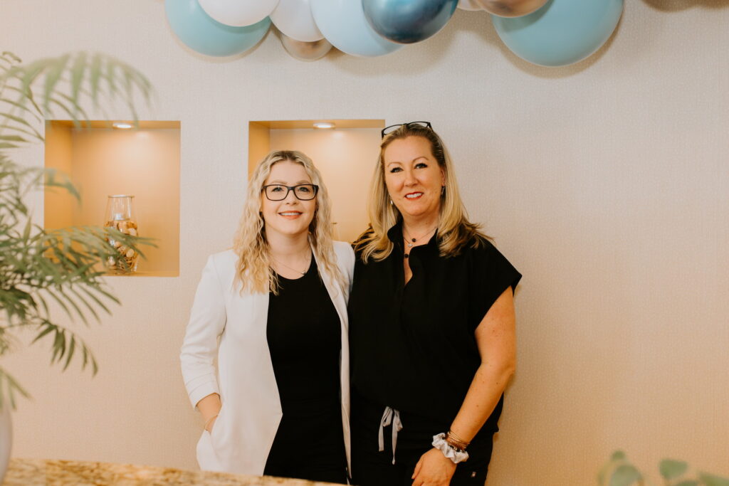 Two smiling women stand together in a welcoming office environment, with a backdrop of decorative balloons in shades of blue and silver. The scene conveys friendliness and professionalism, perfect for a dental practice. The 123Dentist Smiley can be seen nearby, adding a cheerful touch.