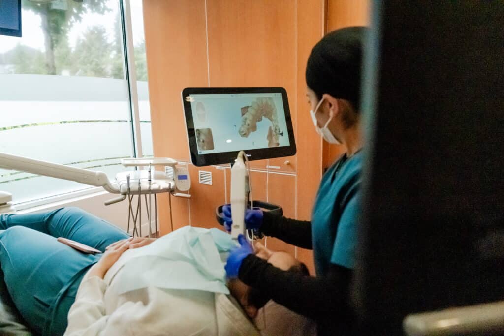 A dental professional in scrubs uses equipment on a patient reclining in a dental chair. A screen displays dental imagery while the environment is well-lit and modern, emphasizing a comfortable dental experience. The welcoming 123Dentist Smiley adds a friendly touch to the scene.