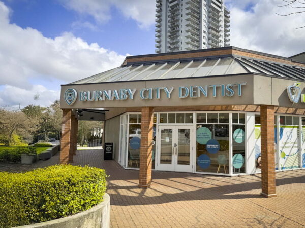 A modern dental clinic named Burnaby City Dentist, featuring large windows and a welcoming entrance, is surrounded by greenery and located near a high-rise building under a partly cloudy sky.