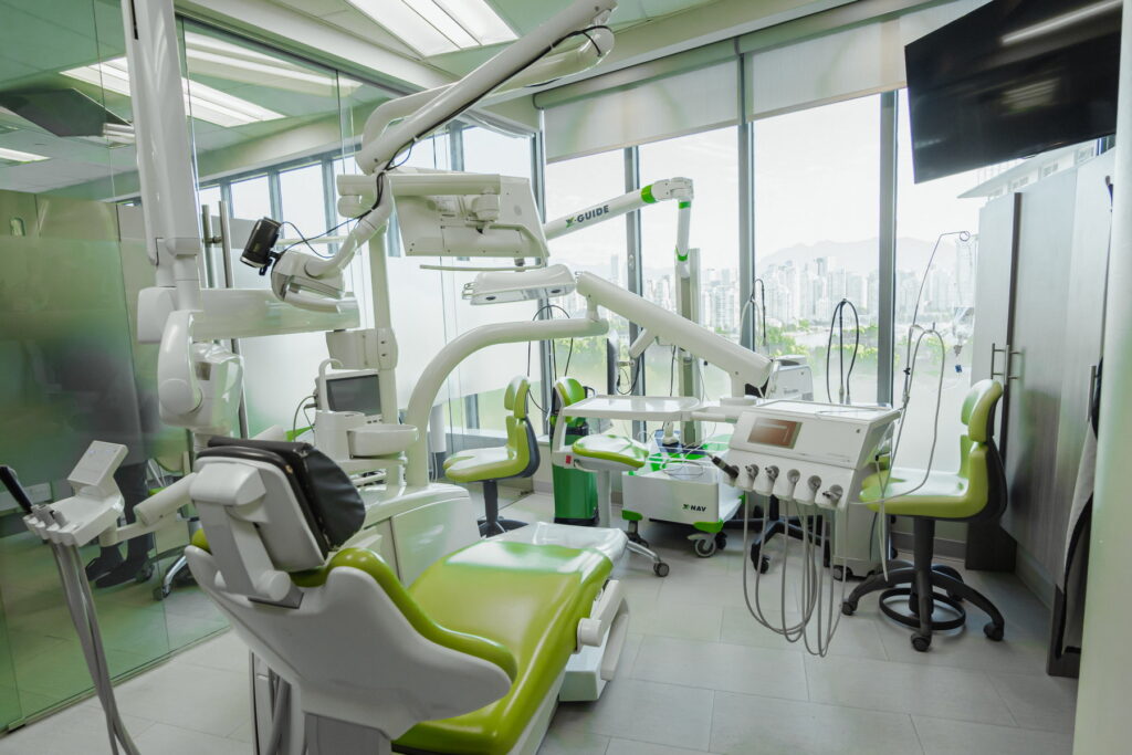 A modern dental clinic interior featuring green and white dental chairs, high-tech equipment, and large windows allowing natural light. The space is clean and organized, creating a welcoming environment for patients.