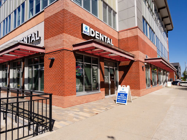 A modern dental office features a red brick exterior with large windows. Signage indicates it is a dental practice. A sidewalk sign points towards services offered, while nearby landscaping adds to the inviting ambiance of the building, offering a friendly atmosphere for visitors.