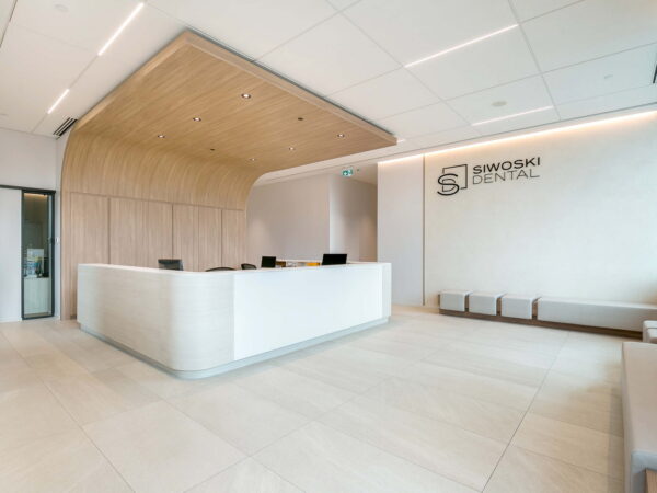 A modern dental office reception area featuring a sleek, white front desk, warm wood accents on the ceiling, and minimalist decor. Natural light illuminates the spacious interior, creating a welcoming atmosphere with a few seating areas visible.