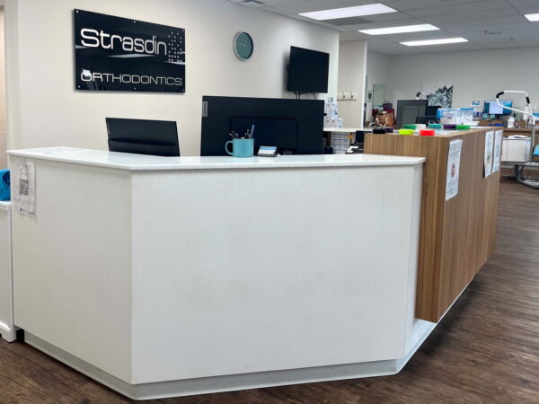 A modern orthodontic office reception area featuring a sleek white desk, a computer, and decorative elements. The space is bright with wooden flooring, and has a welcoming ambiance with chairs and medical equipment visible in the background.