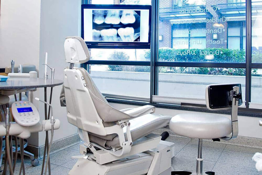 A modern dental office featuring a comfortable dental chair, a monitor displaying dental x-rays, and a bright window offering a view outside. The space is well-lit and clean, creating a welcoming environment for patients. The 123Dentist Smiley adds a friendly touch to the atmosphere.