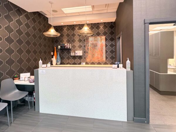 A modern reception area featuring a sleek white desk, contemporary chairs, and stylish pendant lighting. The walls are adorned with a geometric pattern, creating a welcoming atmosphere for visitors. A glimpse of a clinical space is visible through an open doorway.