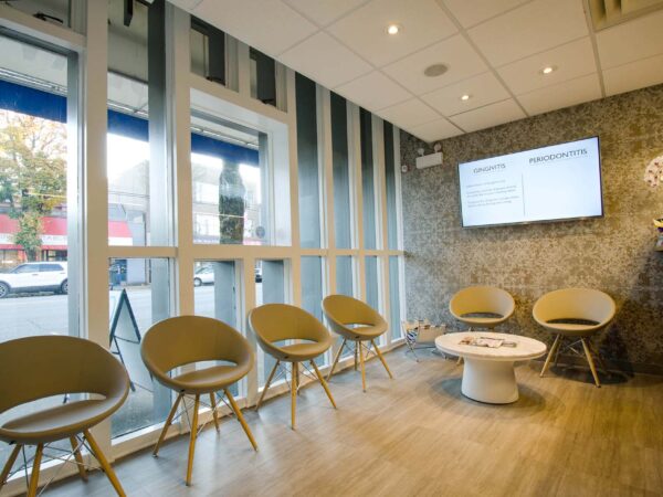 A modern waiting area featuring several beige chairs arranged in a row, a round coffee table in the center, and a large screen displaying information. Natural light streams in through large windows, creating a welcoming atmosphere. The walls are decorated with a minimalist design.