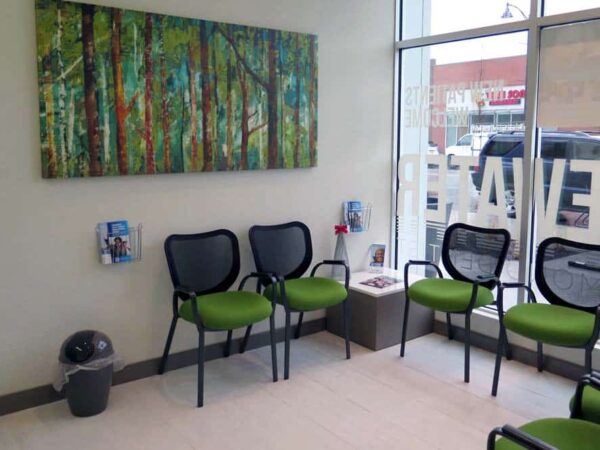 A brightly lit waiting area featuring several green chairs, a trash can, and a wall adorned with a colorful forest painting. A few informational brochures are visible on a small table.