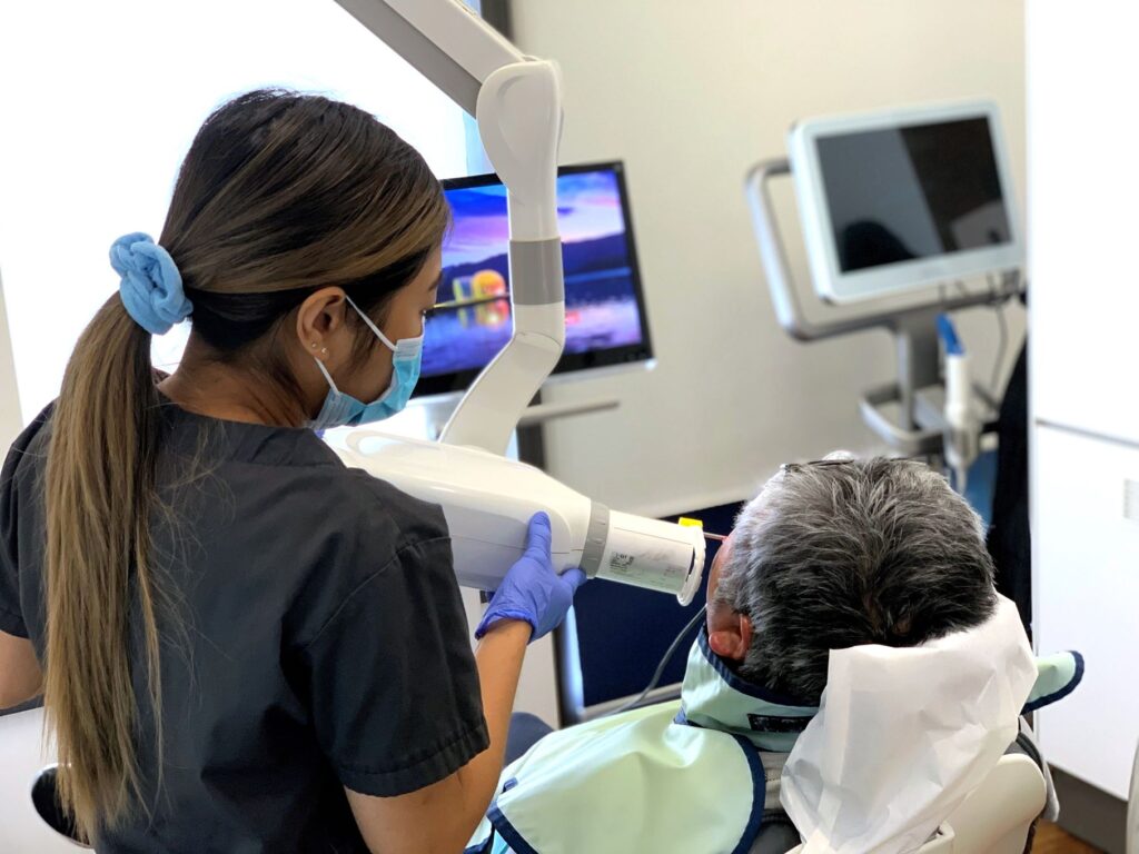 A dental professional in scrubs assists a patient seated in a dental chair, preparing to use a dental scanner. The patient is relaxed with a dental bib on, while monitors display dental images in the background.