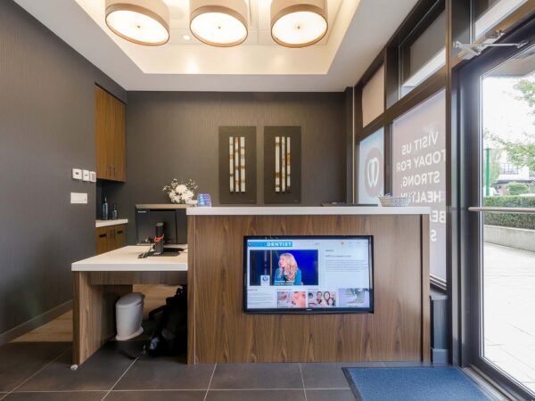 A modern reception area featuring a wooden desk with a screen displaying information, set against a dark wall. Bright overhead lighting and large windows allow natural light in, creating an inviting atmosphere. A small floral arrangement adds a touch of decoration on the counter.