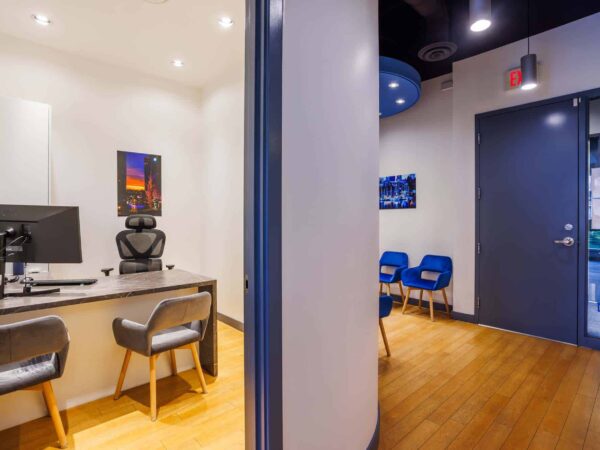 An office interior featuring a desk with a computer, a black ergonomic chair, and framed artwork on the walls. In the background, a waiting area with blue chairs is visible through an open doorway. The space has modern lighting and wooden flooring.