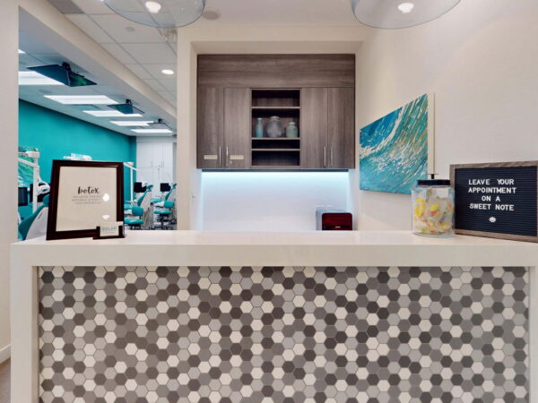 A modern reception desk with a honeycomb-patterned surface, surrounded by a bright, teal wall. The area features a piece of artwork and a small chalkboard sign. Shelving above displays decorative items, creating a welcoming atmosphere.