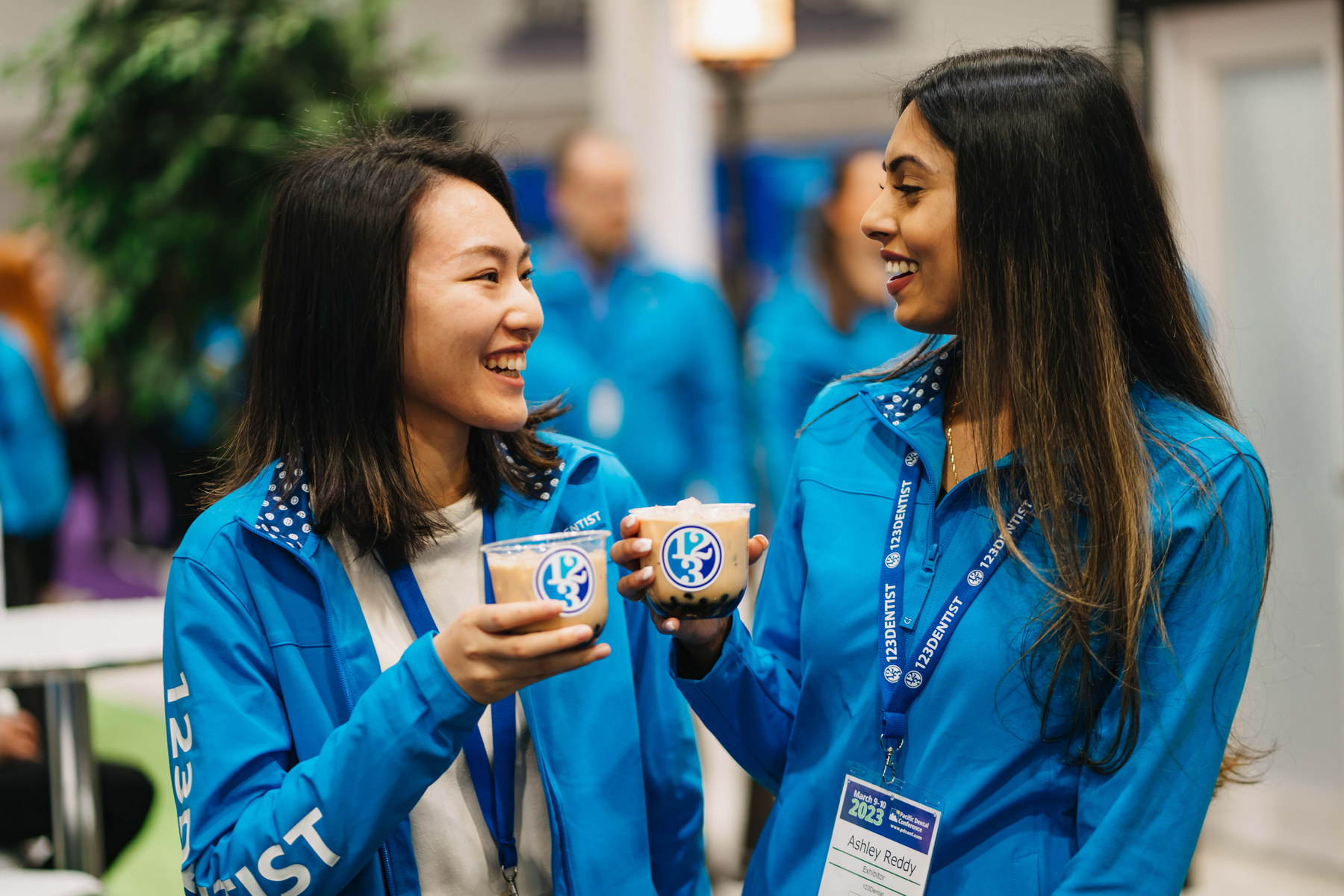 Two women wearing blue jackets smile and hold cups with the 123Dentist logo, engaged in a friendly conversation at an event. In the background, others in blue attire can be seen, creating a lively atmosphere.