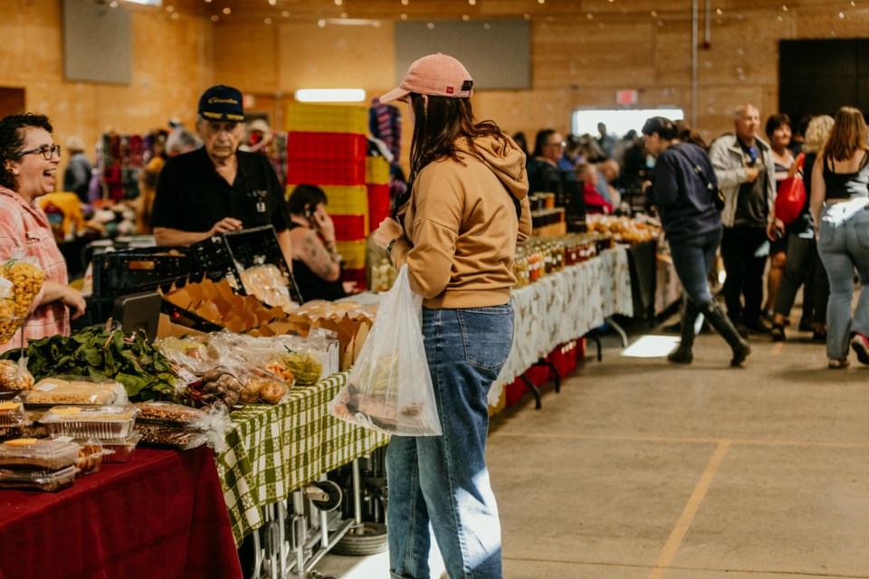 A busy indoor market scene featuring shoppers browsing various stands. A person in casual clothing holds a shopping bag while inspecting items on a table. Vendors interact with customers in a vibrant, bustling atmosphere filled with colorful displays of food and goods.
