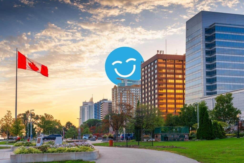 Une vue vibrante du coucher de soleil sur l'horizon d'une ville avec des bâtiments modernes, un drapeau canadien et une icône bleue souriante en superposition, mettant en évidence un sentiment de positivité dans un cadre de parc public.