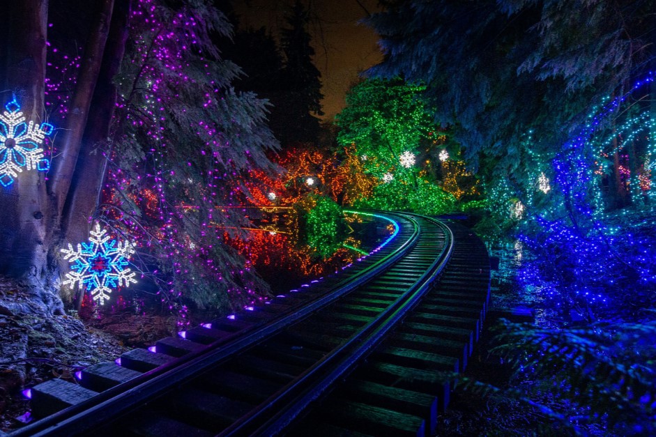 A winding train track illuminated by colorful holiday lights, surrounded by trees. Snowflake decorations hang amidst the vibrant colors, creating a festive night scene.