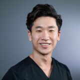 A smiling individual in a black scrub top poses against a neutral gray background, exuding a friendly and approachable demeanor.
