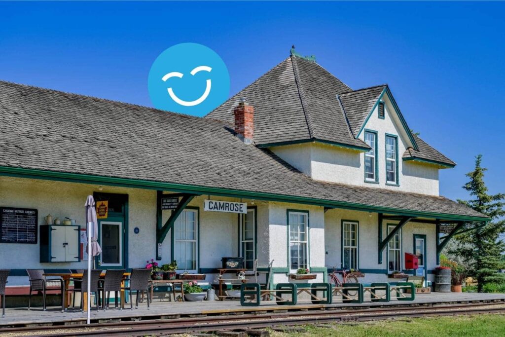 A historic train station with a sloped roof and wooden platform under a clear blue sky. A large blue circle with a smiling face is above the building.