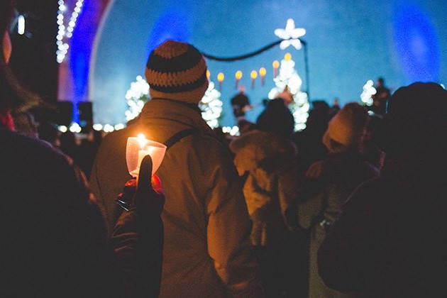 A crowd of people in winter clothing, holding candles, gathered at a festive event with illuminated trees and decorations in the background. A stage with a star-shaped decoration is visible, creating a warm, joyful atmosphere.