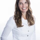 A smiling woman with long, wavy hair wearing a white coat stands against a light background. She appears friendly and approachable, suggesting a professional or healthcare setting.